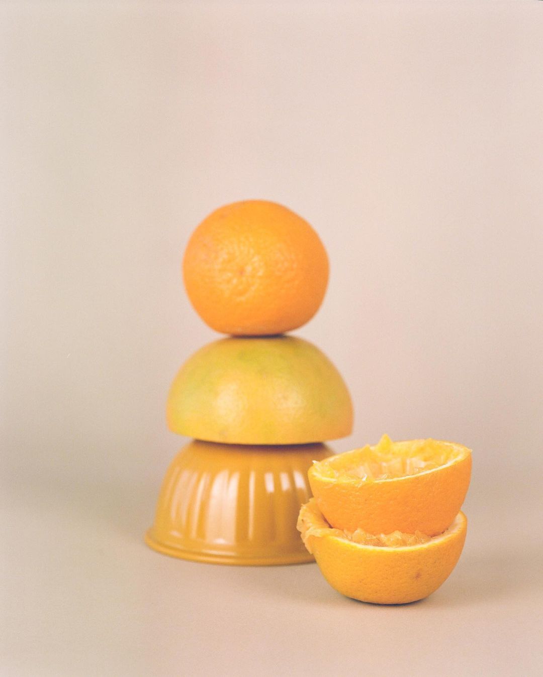 Still life photo of stacked fruit and bowls on a neutral surface, with an orange on top, a yellow fruit, two orange halved fruit and bowls.