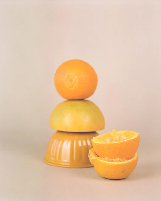 Still life photo of stacked fruit and bowls on a neutral surface, with an orange on top, a yellow fruit, two orange halved fruit and bowls.