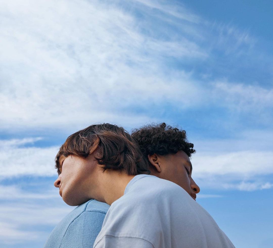 Two people stand back to back against a partly cloudy blue sky, both with their eyes closed, in a portrait-style shot.