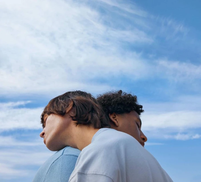 Two people stand back to back against a partly cloudy blue sky, both with their eyes closed, in a portrait-style shot.