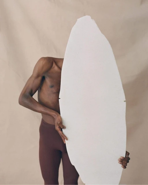 An athletic Black male in maroon leggings stands against a beige backdrop, partially concealed by a large, irregular white stone slab.