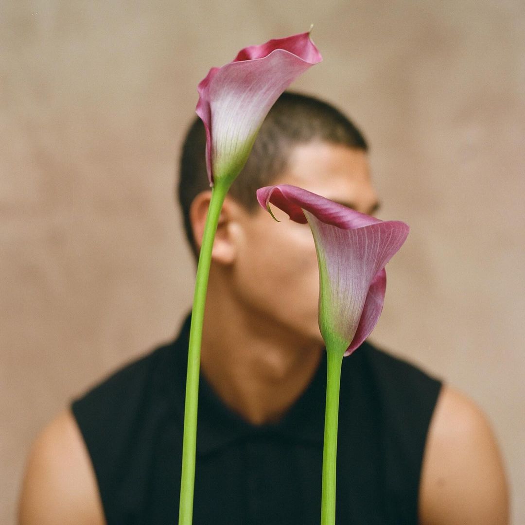 Man partially obscured by calla lilies in a studio setting, wearing a black tank top. The flowers are pink and white with green stems.