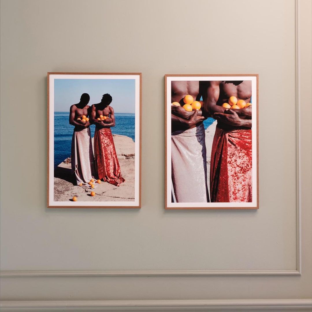 Two framed art prints featuring topless, dark-skinned men holding oranges and wearing shiny skirts, are mounted on a light green wall.
