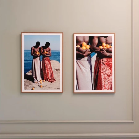 Two framed art prints featuring topless, dark-skinned men holding oranges and wearing shiny skirts, are mounted on a light green wall.