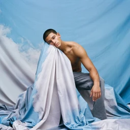 Shirtless man resting his head against draped blue and white cloud-patterned fabric wearing grey trousers