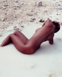 Black woman reclining in shallow milky white water against a rocky, sandy bank
