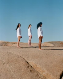 Three women standing barefoot on large smooth rock formations under a clear blue sky, wearing oversized white shirts.