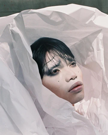 Portrait of a person with dark hair and dramatic eye makeup framed by crumpled white paper structures.