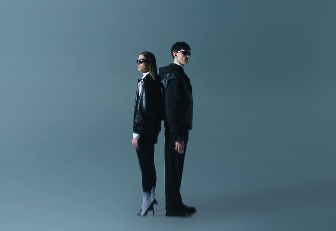 Two people stand back to back in a photo studio. A woman in heels and a man in a jacket pose against a solid blue background.