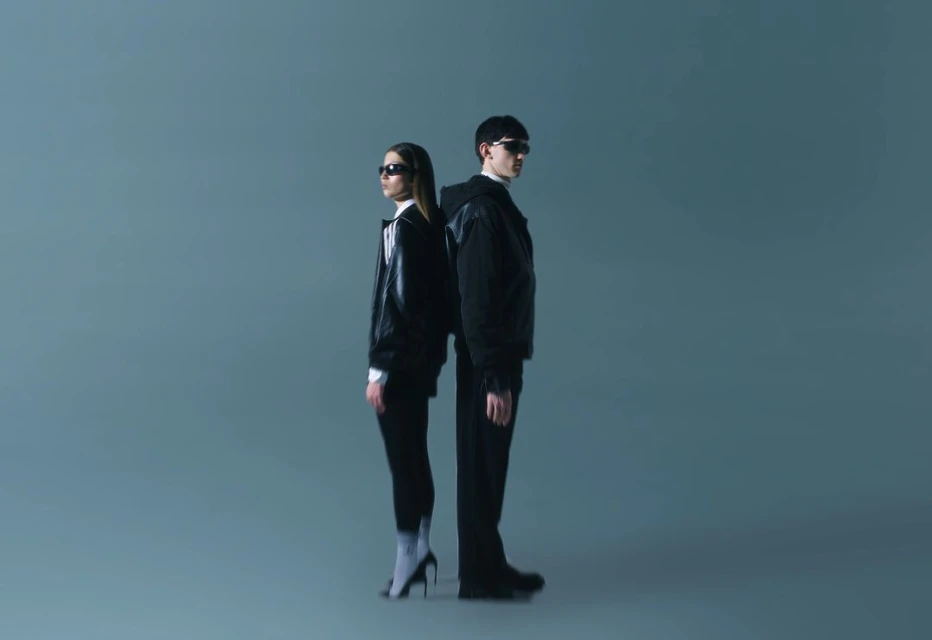 Two people stand back to back in a photo studio. A woman in heels and a man in a jacket pose against a solid blue background.