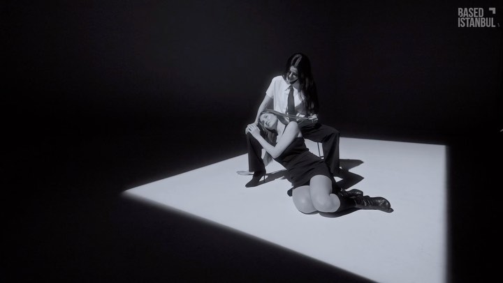 Two women in black and white: one seated in business attire, the other reclined at her feet in a black dress and boots, against stark backdrop.