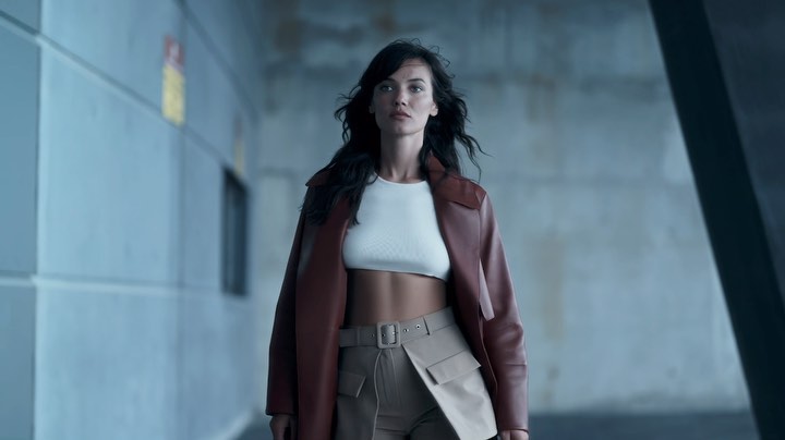 Woman wearing a brown leather coat, white top, and belted trousers standing before a concrete wall
