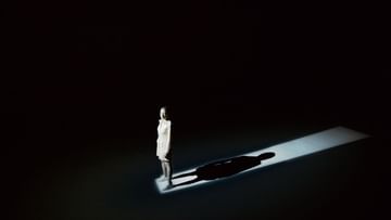Woman in bright light stands in the dark with shadow behind her. Strong lighting creates contrast. Solitary figure.