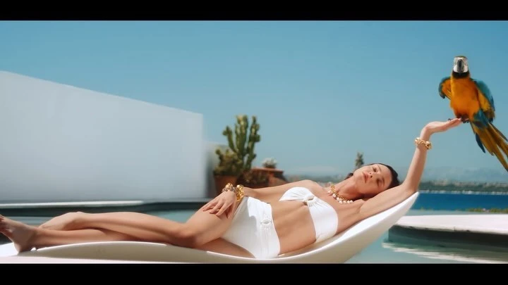Woman in a white bikini lounges by a pool with a macaw perched on her hand, set against a backdrop of blue sea and sky.