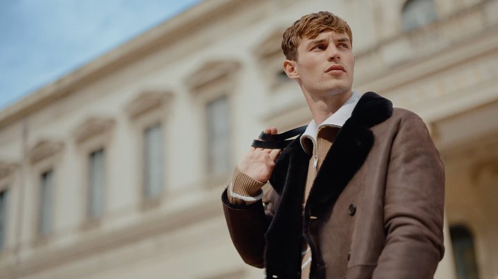 Man in brown shearling coat, outdoors in front of a building, looking off-camera with straps over his shoulder in a low-angle shot.