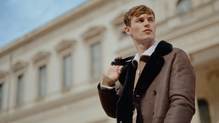 Man in brown shearling coat, outdoors in front of a building, looking off-camera with straps over his shoulder in a low-angle shot.