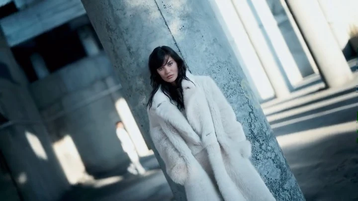 A woman in a white fur coat poses against a concrete column, with another person visible in the background, shot in an urban setting.