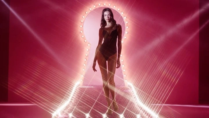 Adult woman in dark bodysuit walks through keyhole-shaped frame with lights in a studio setting with pink background and geometric patterns.