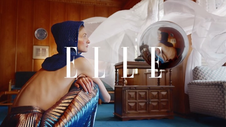 An adult woman in a blue knitted hood poses inside with a vintage cabinet, floating ball, and fashion magazine logo, ELLE.