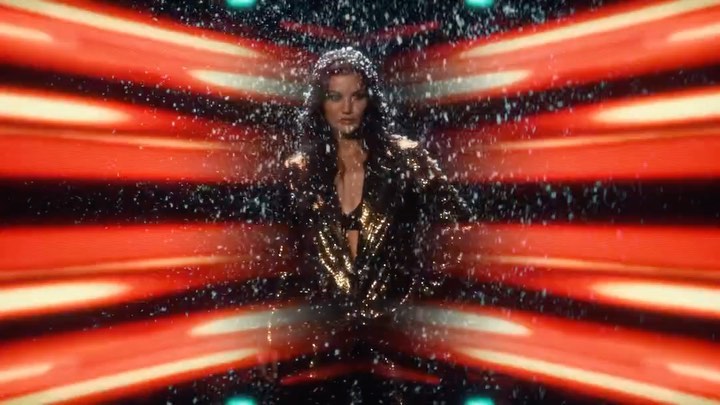 A woman in a glittering gold jacket poses against a dynamic red and black background with white particles falling around her.
