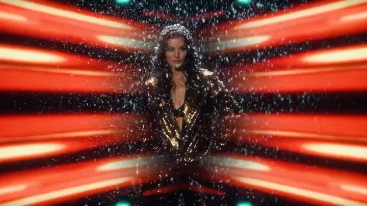 A woman in a glittering gold jacket poses against a dynamic red and black background with white particles falling around her.