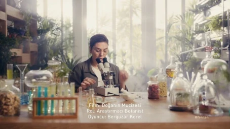Woman in a laboratory setting looking through a microscope surrounded by scientific glassware and plants.