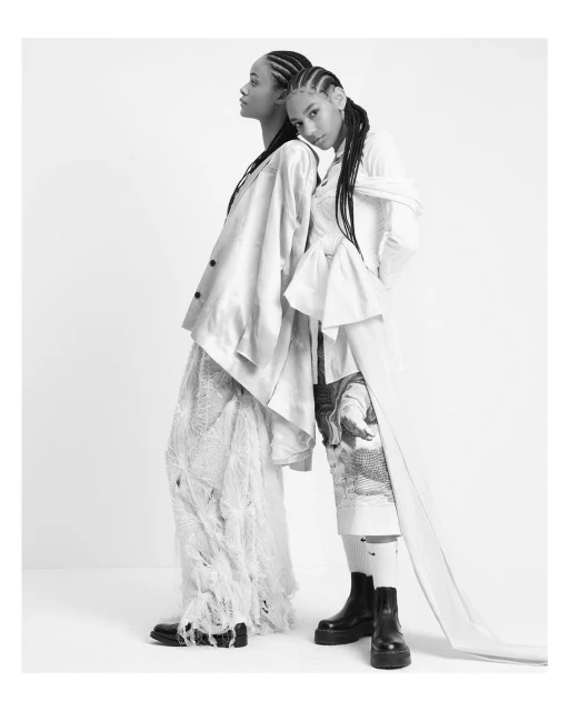 Two models in a black-and-white studio shot wearing draped fabric, pants, and chunky platform boots against a stark white background.