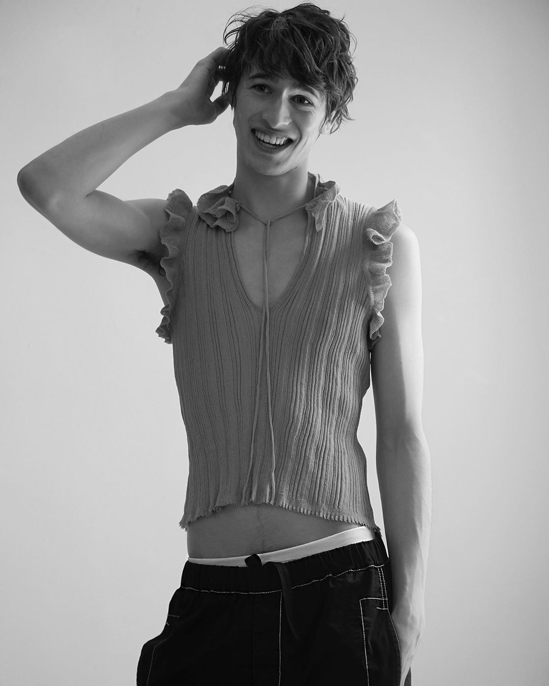 Monochrome portrait of a young adult male with dark hair, wearing a ruffled top and dark pants, hand on head, smiling against a white background.