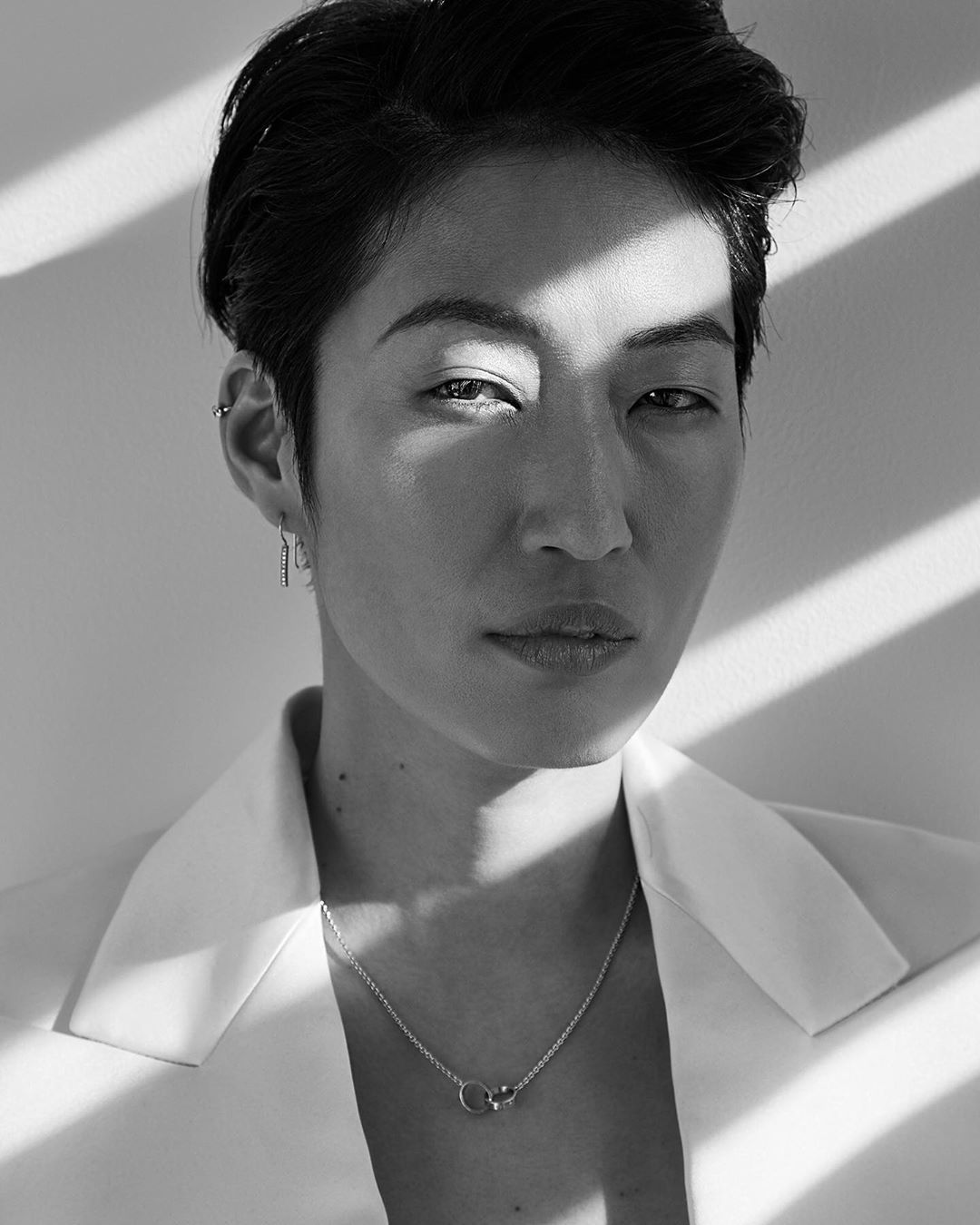 Portrait of a stylish adult woman with short dark hair wearing a white suit jacket and silver necklace, with window shadow pattern background.