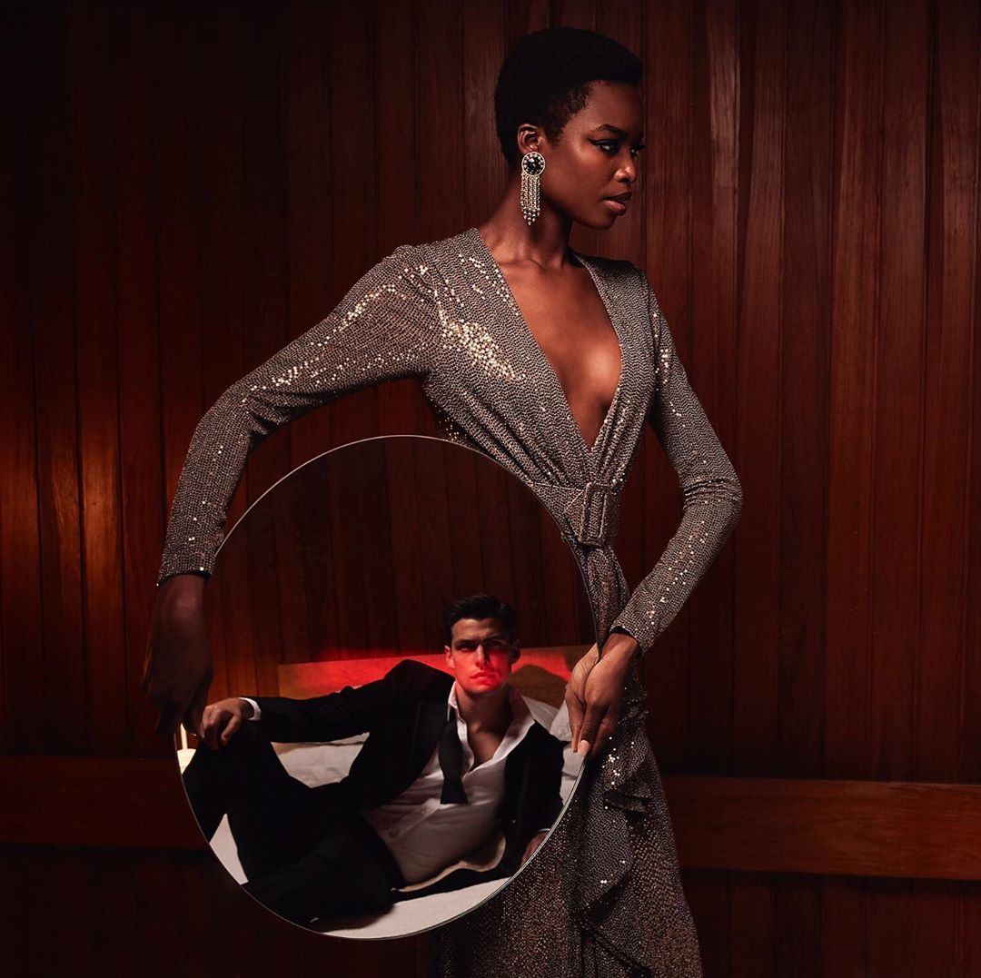 An adult Black woman in a silver dress holds a round mirror reflecting a man in a suit, against a dark wood-panelled background.