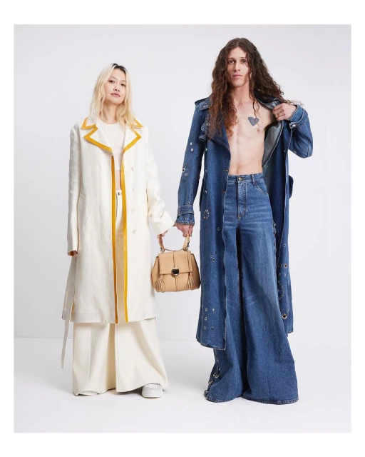 Two fashion models pose in a photo studio wearing long denim and linen trench coats with matching wide-leg pants and a tan leather handbag.