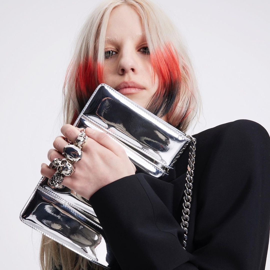 Close-up of a fair-skinned adult woman holding a reflective silver clutch purse with chain, wearing a black jacket in a studio.