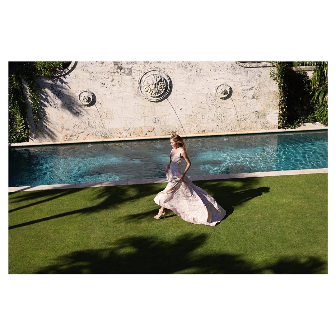 Woman in elegant gown walking along pool in lush outdoor setting with historic stone architecture and green grass. Fashion editorial shot.
