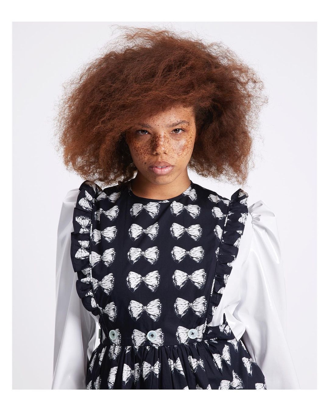 Woman with freckles poses in a studio, wearing a black and white bow-patterned dress with white sleeves and ruffled trim, against a white background.