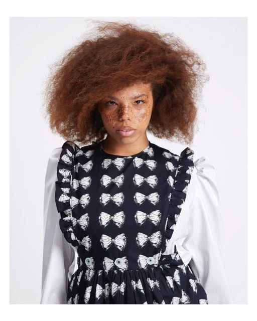 Woman with freckles poses in a studio, wearing a black and white bow-patterned dress with white sleeves and ruffled trim, against a white background.