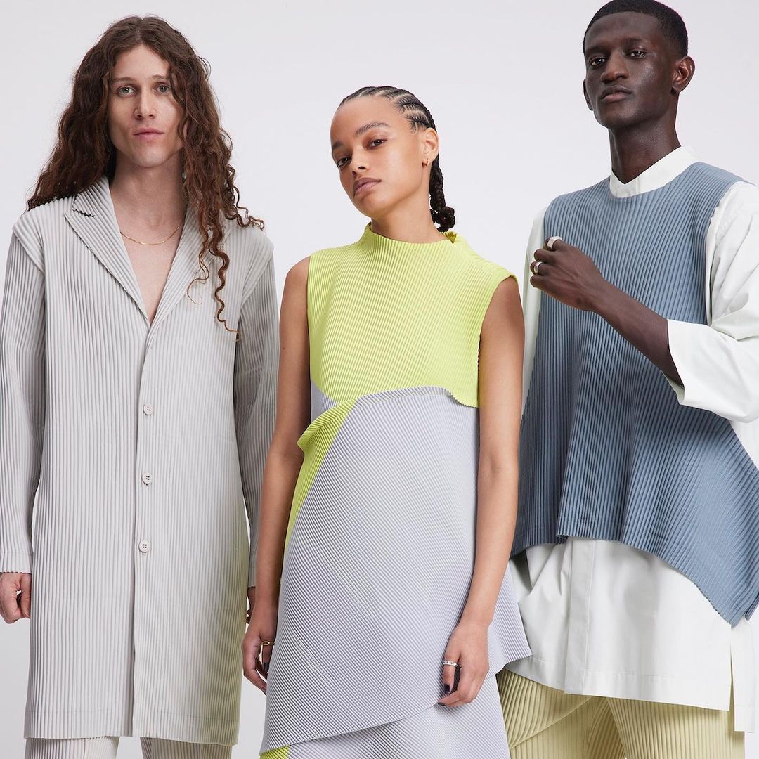 Three fashion models in a studio wearing colorful pleated dresses in a fashion editorial shot on a bright white background with studio lighting.