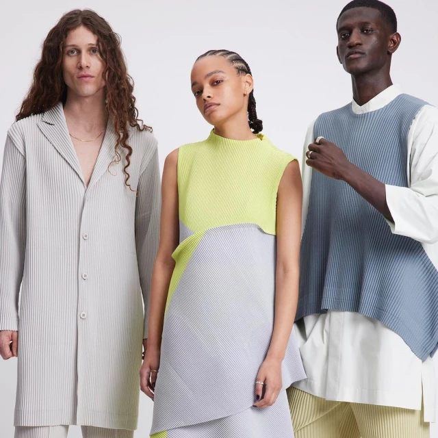 Three fashion models in a studio wearing colorful pleated dresses in a fashion editorial shot on a bright white background with studio lighting.