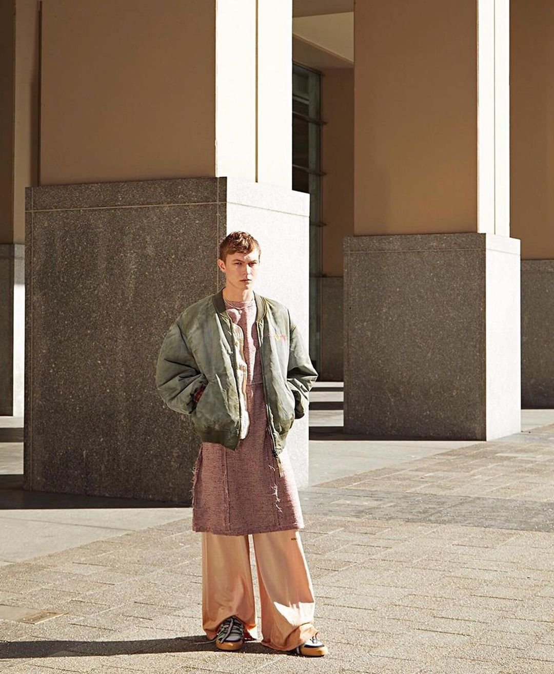 Young adult male in a green jacket and peach colored pants standing in an urban outdoor setting with concrete columns.