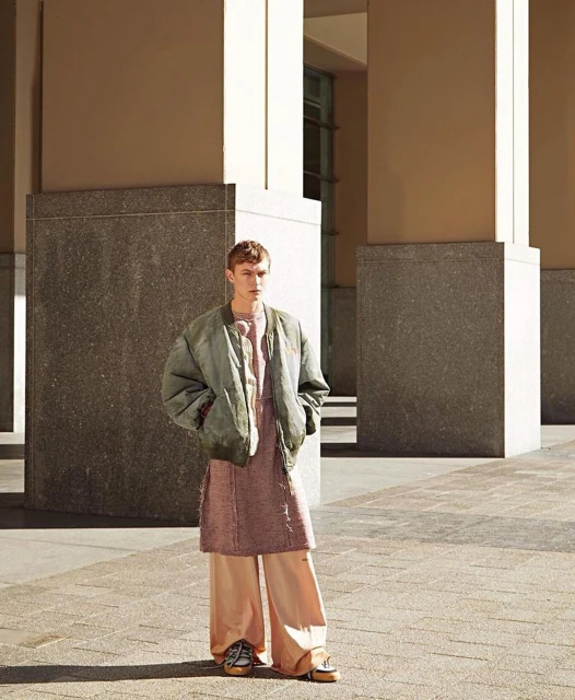 Young adult male in a green jacket and peach colored pants standing in an urban outdoor setting with concrete columns.