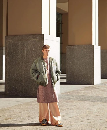 Young adult male in a green jacket and peach colored pants standing in an urban outdoor setting with concrete columns.