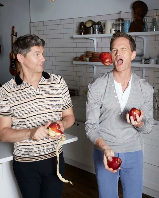 Two adult males in a kitchen: one peels a red apple, while the other juggles and winks, with white tile and cabinets in the background.