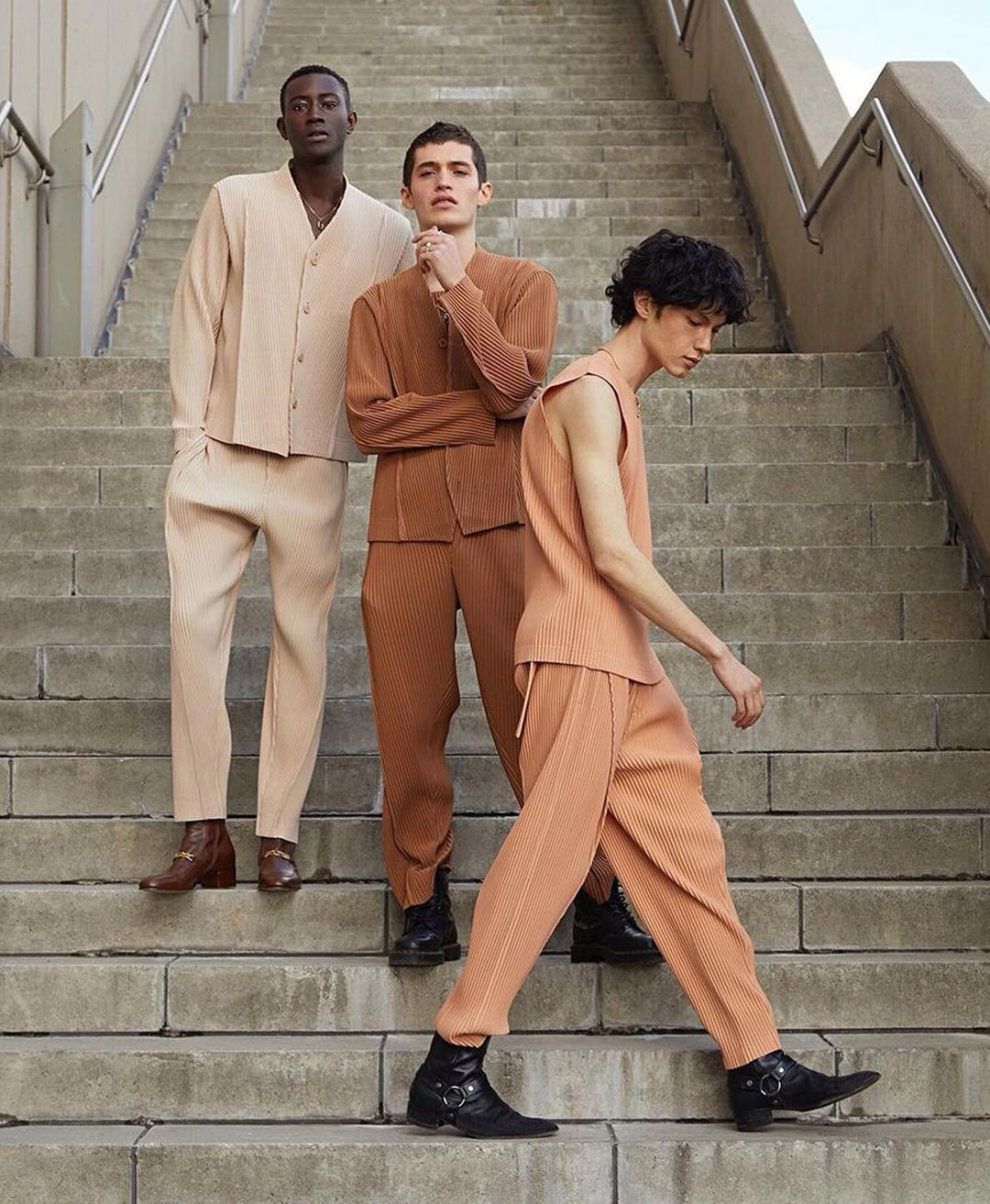 Three male fashion models pose on outdoor concrete stairs wearing complementary-colored pleated ensembles and boots in a lifestyle fashion shoot.