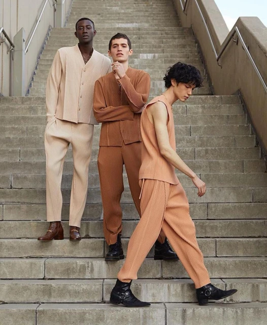 Three male fashion models pose on outdoor concrete stairs wearing complementary-colored pleated ensembles and boots in a lifestyle fashion shoot.