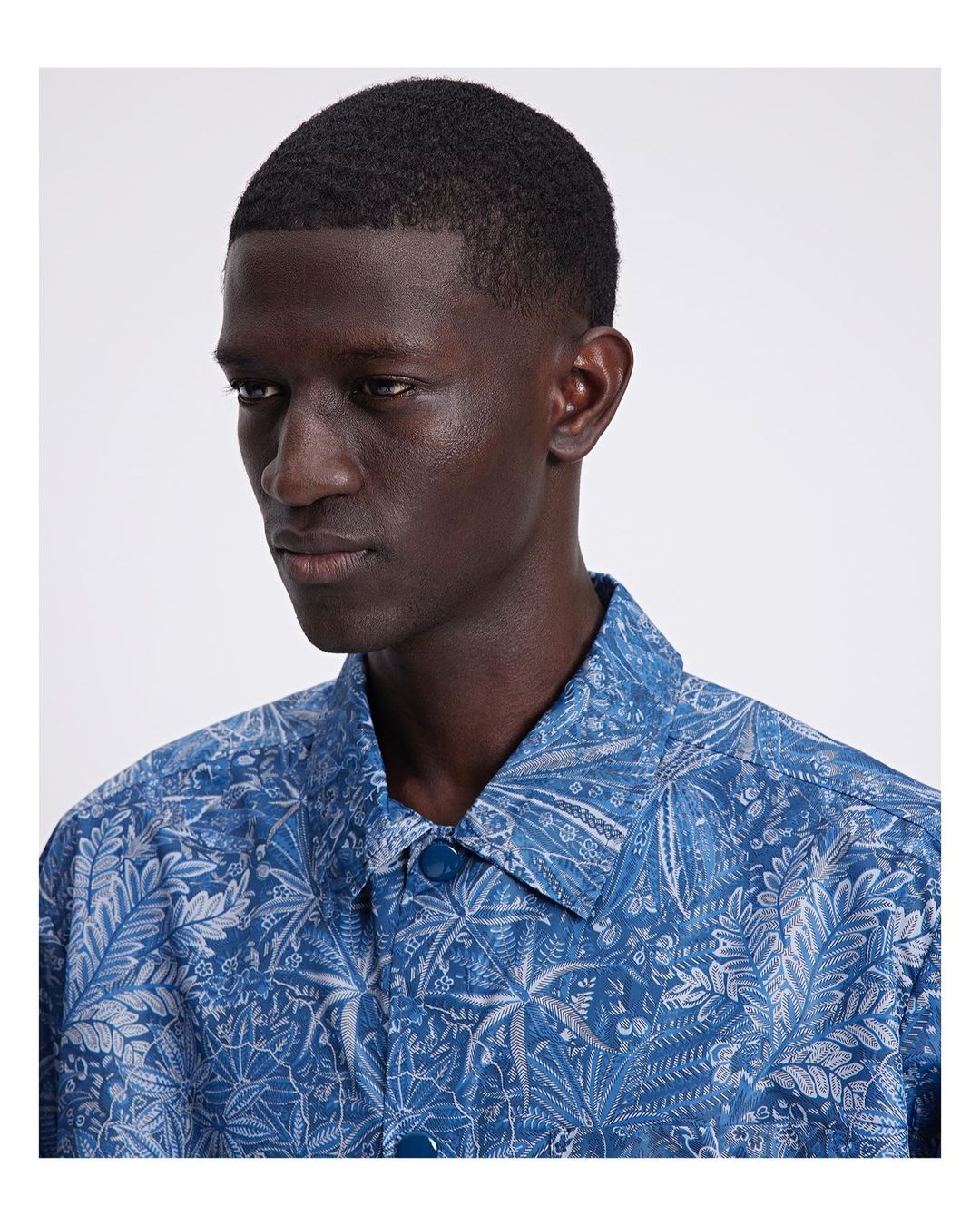 Close-up of a dark-skinned adult male wearing a blue collared shirt with a light gray floral print, facing slightly left.
