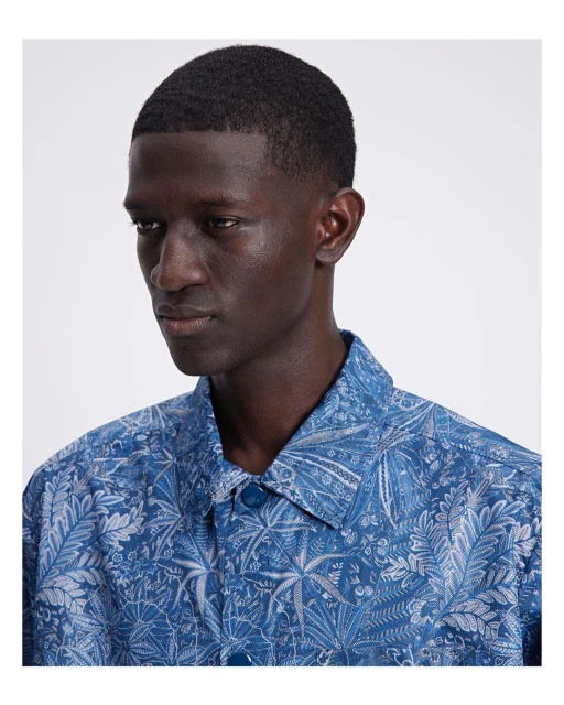 Close-up of a dark-skinned adult male wearing a blue collared shirt with a light gray floral print, facing slightly left.