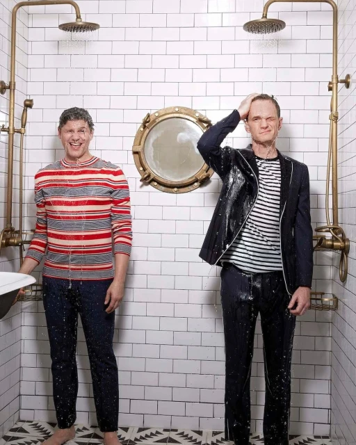 Two adult men in a tiled shower with running water, one in a striped top, the other in a suit jacket, under brass shower heads.