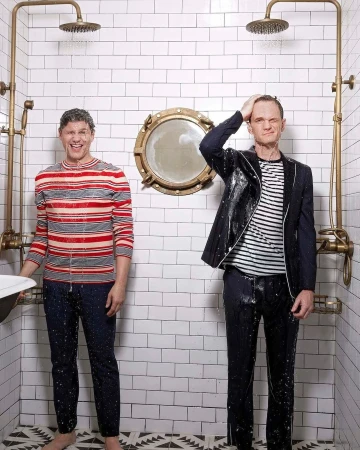 Two adult men in a tiled shower with running water, one in a striped top, the other in a suit jacket, under brass shower heads.