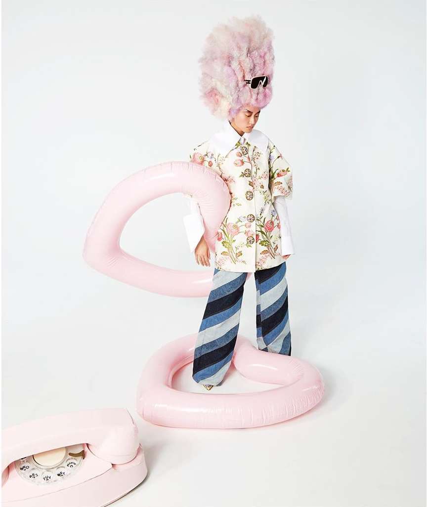 Child wearing floral jacket with striped pants and a pink wig, standing next to pink inflatable objects in a white photo studio.