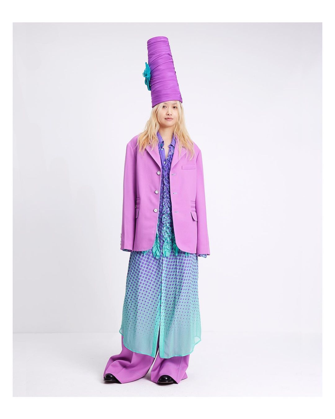 A full-length portrait of a woman wearing a purple suit, tall hat, and floral-print dress, posing against a white studio background.