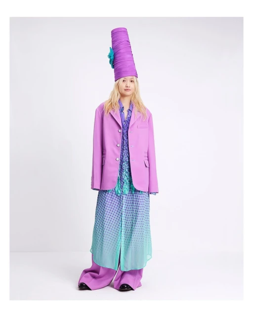 A full-length portrait of a woman wearing a purple suit, tall hat, and floral-print dress, posing against a white studio background.